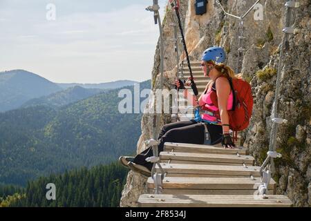Woman tourist admires the view sitting down on a via ferrata suspended bridge at Lacul Rosu, Neamt county, Romania, on a route called Wild Ferenc. Stock Photo