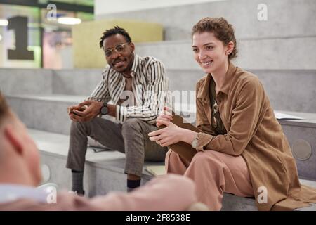 Group of students chatting cheerfully while relaxing in modern school lounge, focus on smiling young woman talking to friend Stock Photo