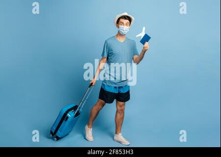 Full length caucasian joyful tourist in a hat and medical mask, holds a blue suitcase and a small airplane with passport, looks at camera, happily smiling, stands on an isolated blue background Stock Photo