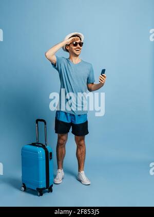 Happy caucasian male tourist, in a hat and sunglasses, looks into the distance to meet adventures, next to him a blue suitcase, rejoices a long-awaited vacation, stands on an isolated blue background Stock Photo