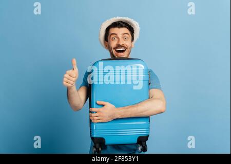 Amazed joyful caucasian male tourist, in a summer hat, tightly hugs a blue suitcase, shows thumbs up gesture, smiles happily, stands on an isolated blue background Stock Photo