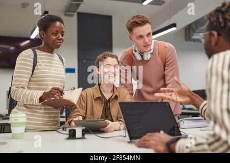 Diverse group of students discussing project at desk while working together at school library and smiling cheerfully Stock Photo