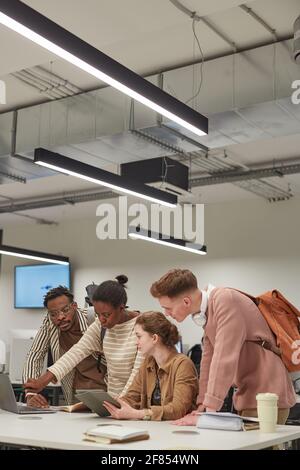 Vertical shot of diverse group of students working together and using digital tablet while standing at table in school IT lab, copy space Stock Photo