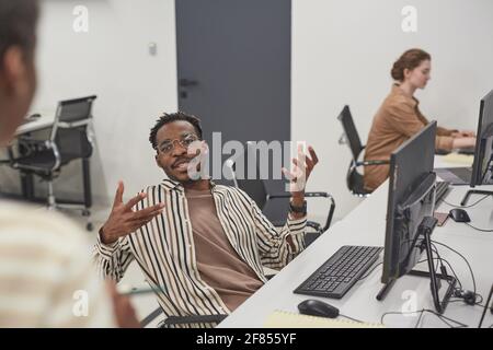 Portrait of African-American man talking to colleague in office and gesturing actively while sitting at workplace with computer, copy space Stock Photo