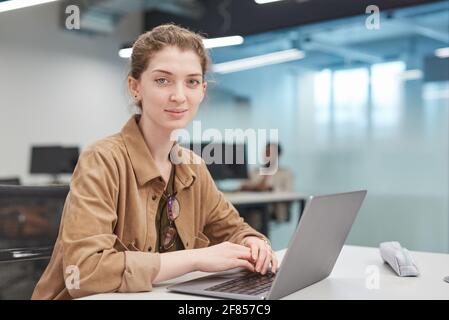 Portrait of young woman using laptop at smiling at camera in office or school library setting, copy space Stock Photo