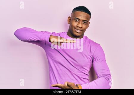 Young black man wearing casual pink sweater gesturing with hands showing big and large size sign, measure symbol. smiling looking at the camera. measu Stock Photo