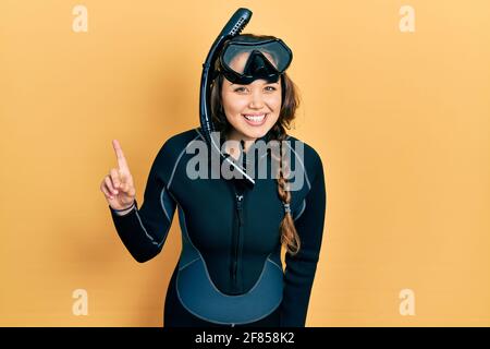 Young hispanic girl wearing diver neoprene uniform smiling with an idea or question pointing finger up with happy face, number one Stock Photo