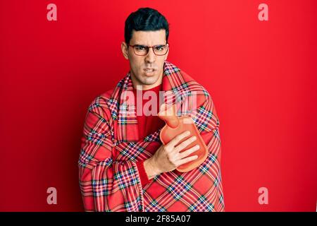 Handsome hispanic man wrapped in a winter blanket holding hot water bag clueless and confused expression. doubt concept. Stock Photo
