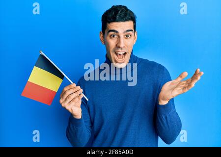 Handsome hispanic man holding belgium flag celebrating achievement with happy smile and winner expression with raised hand Stock Photo