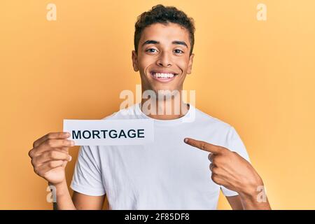 Young handsome african american man holding mortgage word smiling happy pointing with hand and finger Stock Photo