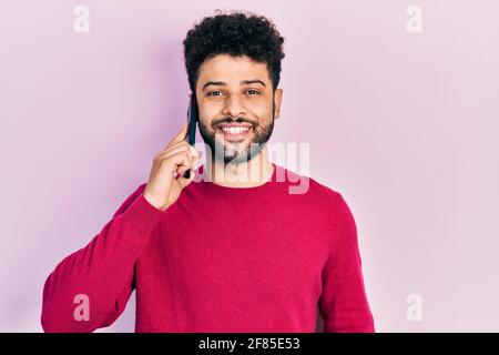 Young arab man with beard having conversation talking on the smartphone looking positive and happy standing and smiling with a confident smile showing Stock Photo
