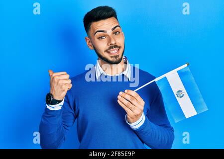 Young hispanic man with beard holding el salvador flag pointing thumb up to the side smiling happy with open mouth Stock Photo