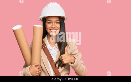 Beautiful hispanic woman holding paper blueprints smiling happy and positive, thumb up doing excellent and approval sign Stock Photo
