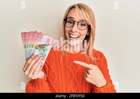 Beautiful blonde woman holding 100 new zealand dollars banknote smiling happy pointing with hand and finger Stock Photo