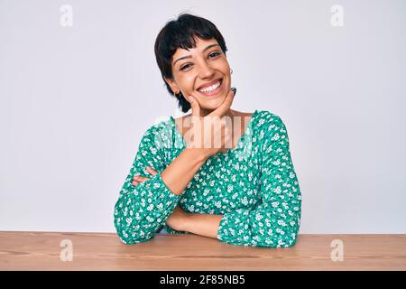 Beautiful brunettte woman wearing casual clothes sitting on the table looking confident at the camera smiling with crossed arms and hand raised on chi Stock Photo