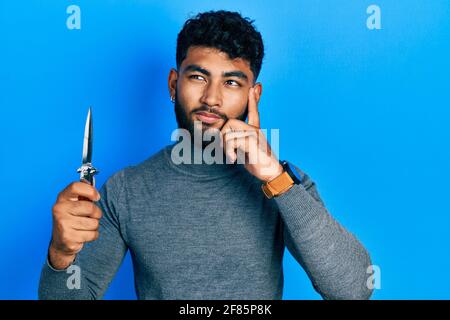 Arab man with beard holding pocket knife serious face thinking about question with hand on chin, thoughtful about confusing idea Stock Photo
