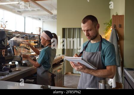 Waist up portrait of male barista wearing apron while working in coffee shop and holding clipboard, copy space Stock Photo