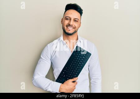 Young arab man holding computer keyboard looking positive and happy standing and smiling with a confident smile showing teeth Stock Photo