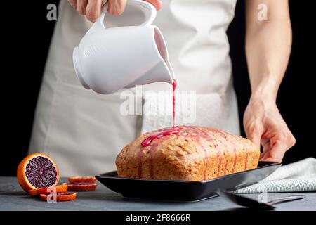 Closeup image of fresh homemade blood orange cake made in a loaf pan and served on black porcelain plate on dark stone background.  A woman pours bloo Stock Photo