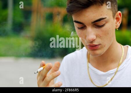 Young man with pimply skin, gold earring and matching chain smoking a cigarette outdoors exhaling a puff of smoke as he looks down with a serious expr Stock Photo