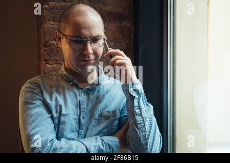 Man in a blue shirt sitting near the window and talking on smartphone. Working at home or modern coworking office. Stock Photo