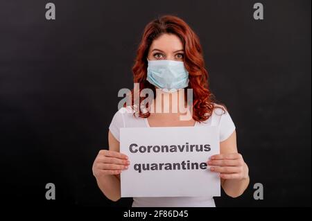 Frightened woman in a medical mask holds a poster. Red-haired girl on black background in an antimicrobial quarantine respirator. Coronavirus COVID-19 Stock Photo
