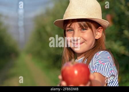 Portrait Of Smiling Little Girl With Blue Eyes in an Orchard. Healthy Food Concept. Close up Face Shot of Cute Girl with Hat. Stock Photo