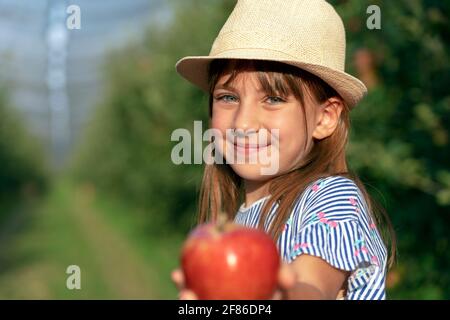 Portrait Of Smiling Little Girl With Beautiful Eyes in an Orchard. Healthy Food Concept. Cute Girl with Hat Holding Red Apple in Hand. Stock Photo