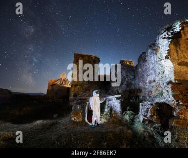 Side view of spaceman in special white suit and helmet research ancient ruins with lantern under starry sky at night. Concept of walking and research. Stock Photo