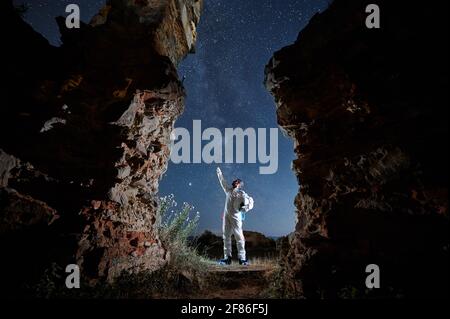 Front view of spaceman in special suit standing and looking on starry sky near ancient ruins at night. Astronaut exploring stars in sky. Stock Photo