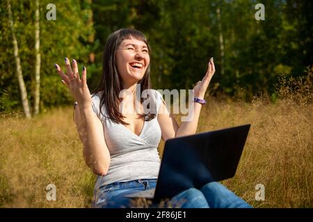 happy young woman with laptop on a sunny lawn. freelancer works in nature. student studies remotely on nature landscape outdoor Stock Photo