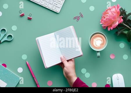Save the date invitation card design. Hand with pen, home office. Text space, copy-space in notebook..Pink peony flowers on green mint table. Coffee, Stock Photo