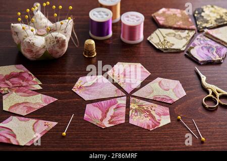 Pentagonal pieces of fabric lying in the shape of a flower, stacks of multi-colored pieces of fabric, sewing accessories on a desk