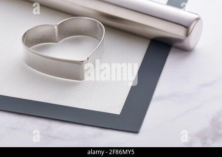 Stainless steel heart-shaped cookie cutter and rolling pin lying on a baking mat Stock Photo