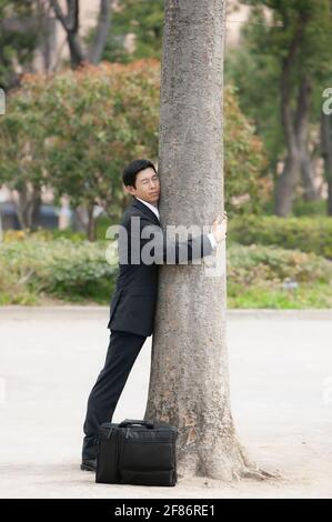 Serene businessman in suit hugging tree in park Stock Photo