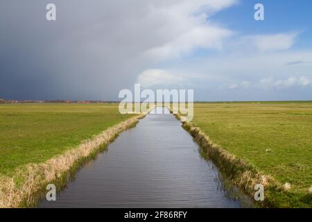 Storm front approaching over rural landscape on the Dutch island Terschelling: dark threatening clouds above a flat polder landscape with a canal Stock Photo