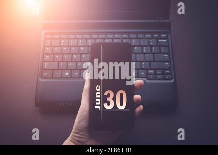 June 30th. Day 30 of month, Calendar date. Hand Holding Mobile Phone on Laptop Computer dark background with sunshine. Summer month, day of the year c Stock Photo