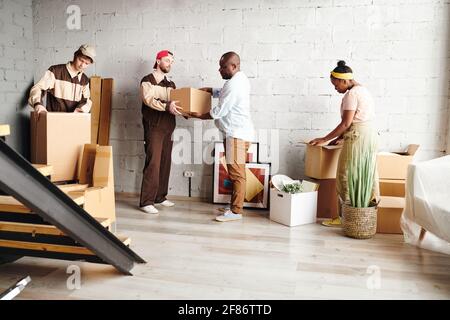 Two young workers in uniform helping married African couple with delivering packed boxes to new flat or house during remove Stock Photo