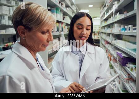 Two female pharmacists discussing in chemist using digital table while standing between aisle Stock Photo