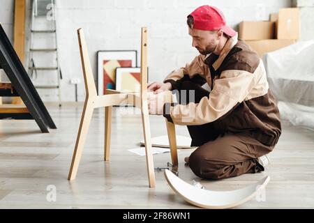 Serious young furniture assemblage specialist in workwear standing on knees on the floor and assembling wooden chair in new flat or studio Stock Photo