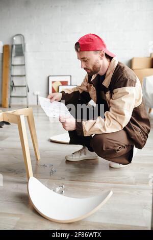 Serious assemblage specialist in workwear standing on squats and looking through instruction on paper while assembling furniture of client Stock Photo