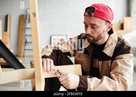 Serious young furniture assemblage specialist in workwear fixing parts of chair while assembling it in large new house, flat or studio Stock Photo