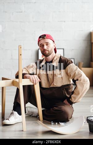Serious young furniture assemblage specialist in workwear sitting on the floor by unfinished chair and looking at you in new flat or studio Stock Photo