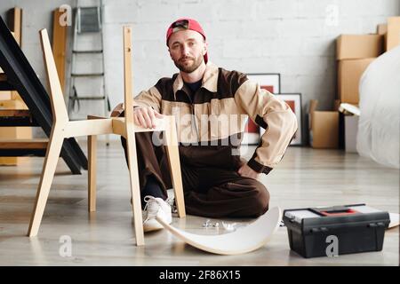 Young specialist in assembling furniture sitting on the floor by unfinished chair in new flat or studio and looking at you while working Stock Photo
