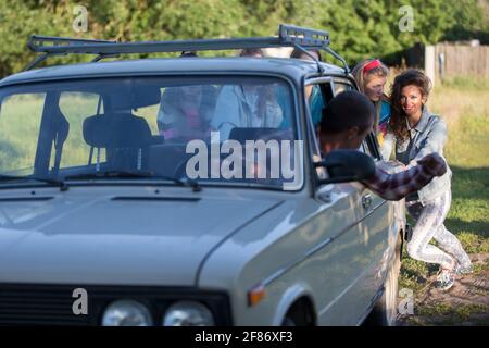 Young cheerful girls are pushing an old car. Women in the style of the 90s. Stock Photo