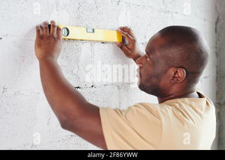 Young man of African ethnicity standing in front of white painted brick wall and using level measuring handtool during home renovation Stock Photo