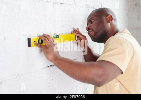 Young man of African ethnicity putting level measuring handtool to white painted brick wall while checking its flatness during home renovation Stock Photo