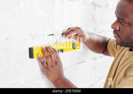 Young man of African ethnicity with pencil drawing line on white painted brick wall while using level measuring handtool during home renovation Stock Photo