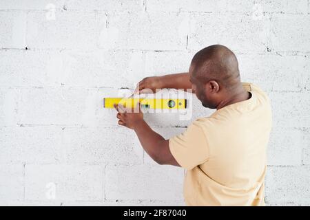 Young African man with pencil drawing line over level measuring handtool on white painted brick wall during renovation of living-room Stock Photo