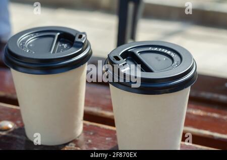 Two paper cups with a lid on a park bench. Close-up of eco-friendly disposable hot drinks takeaway tableware. Daytime. Outdoors. Stock Photo